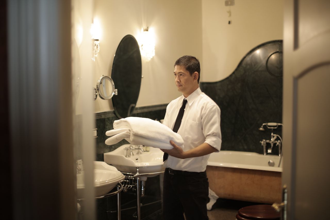 services-02 Vue de côté d’un jeune employé tenant une pile de serviettes blanches dans la salle de bain d’un hôtel de luxe
